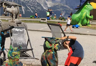 A boy is painting colorful animal figures in an outdoor area. In the background, mountains and more colorful sculptures can be seen.