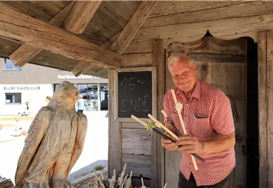 An older man stands in front of a wooden shed, holding some handmade rods in his hand. Next to him, a carved bird sculpture can be seen.