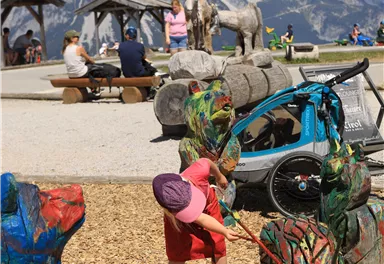 A child is playing on a playground with colorful wooden sculptures. In the background, mountains and various visitors can be seen.