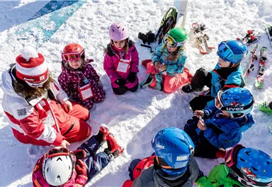 Eine Gruppe von Kindern sitzt im Schnee und hört einem Skilehrer zu. Rundherum sind Skiausrüstungen und verschneite Berge sichtbar.
