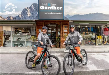 Two cyclists stand with their bicycles in front of a sports store. In the background, the mountains and the shop sign are visible.