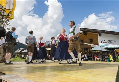 Eine Gruppentanzaufführung im Freien mit traditionell gekleideten Tänzern. Im Hintergrund sind Gäste vor einem Gasthaus auf einer Terrasse zu sehen.