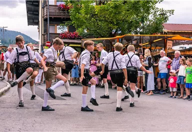 A group of boys is dancing in traditional costumes on a street. People are watching them and enjoying the festive atmosphere.