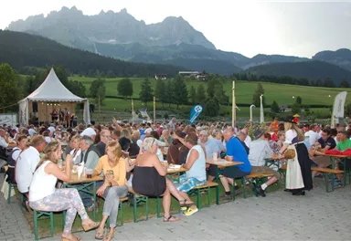 Eine große Menschenmenge sitzt an Biertischen und genießt eine Veranstaltung im Freien. Im Hintergrund sind beeindruckende Berge und ein Zelt zu sehen.