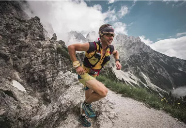 Ein Läufer joggt auf einem schmalen Weg in den Bergen. Die Landschaft ist beeindruckend mit Felsen und Wolken im Hintergrund.