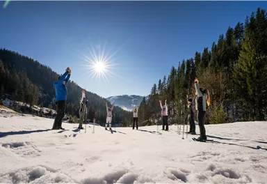 Eine Gruppe von Menschen macht eine Dehnübung im Schnee. Die Sonne scheint hell in den klaren blauen Himmel.