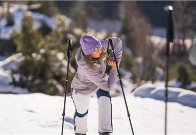 A woman puts on her skiing equipment in a snowy landscape. In the background, trees and a clear winter atmosphere can be seen.