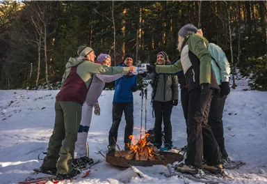 Eine Gruppe von sechs Menschen steht im Schnee um ein Lagerfeuer und stoßt mit Getränken an. Im Hintergrund sind Bäume und eine winterliche Landschaft sichtbar.