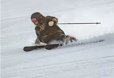 Ein Skifahrer fährt schnell den Hang hinunter und macht eine Kurve im Schnee. Die Umgebung ist weiß und winterlich.