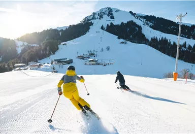 Zwei Skifahrer fahren die Piste hinunter, umgeben von schneebedeckten Bergen. Die Sonne scheint hell am blauen Himmel.