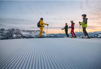 A group of skiers stands on a freshly groomed slope in the mountains. The view shows snow-covered peaks and a clear sky at sunrise.