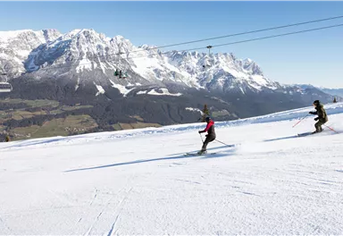 Zwei Skifahrer fahren die schneebedeckte Piste hinunter. Im Hintergrund sind majestätische Berge und ein klarer blauer Himmel zu sehen.