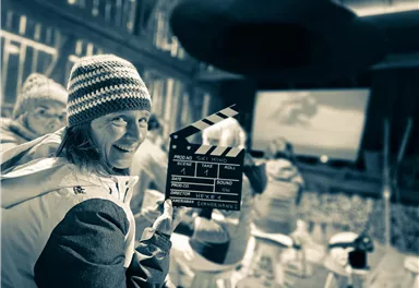 Eine fröhliche Person hält eine Klappe in einem Kino oder Filmraum. Im Hintergrund sind weitere Menschen und eine große Leinwand zu sehen.