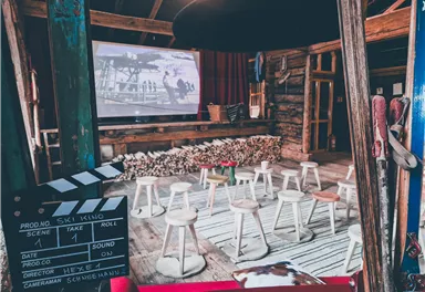 Ein gemütlicher Veranstaltungsraum mit Stühlen und einem großen Bildschirm. Der Raum hat eine rustikale Atmosphäre mit Holzelementen und einer Filmklappe im Vordergrund.