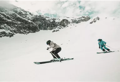 Zwei Skifahrer fahren über einen schneebedeckten Hang. Im Hintergrund sind beeindruckende Berge und ein bewölkter Himmel zu sehen.