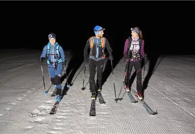 Three skiers are walking at night over a snow-covered slope. They are wearing headlamps and skiing in a deep, dark environment.