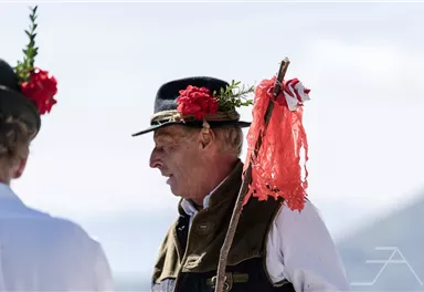 Ein Mann in traditioneller Kleidung mit einer blühenden Kopfbedeckung und einem Stock. Im Hintergrund sind unscharfe Silhouetten von anderen Personen zu sehen.