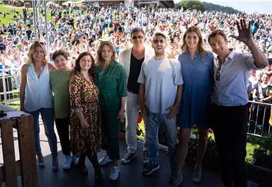 A group of eight people stands smiling together in front of a large crowd. In the background, green meadows and a clear sky can be seen.