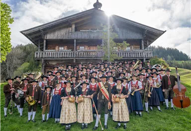 A large group of people in traditional attire gathers in front of a rustic, alpine wooden house. They hold various instruments and stand on a green meadow.