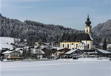A snow-covered landscape with a charming village and a church in the foreground. In the background, snow-capped mountains and a clear sky can be seen.
