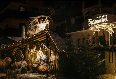 Eine festlich beleuchtete Weihnachtskrippe mit Figuren von Maria, Josef und dem Kind Jesus. Im Hintergrund ist ein geschmücktes Schild mit dem Namen des Ortes sichtbar.