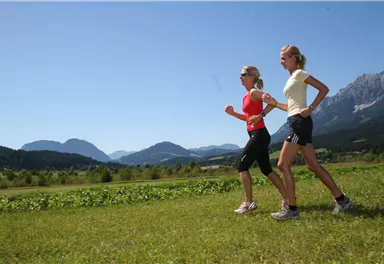 Zwei Frauen joggen auf einem grünen Feld mit Bergen im Hintergrund. Der Himmel ist klar und blau.