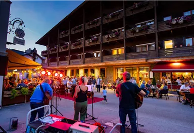 A busy street scene with musicians performing in front of a wooden building. Many people are sitting outside and enjoying the evening atmosphere.