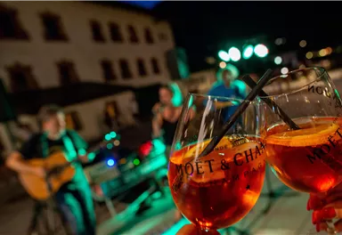 Two glasses with drinks are in the foreground, while a band is playing in the background. The atmosphere is celebratory and cozy at night.