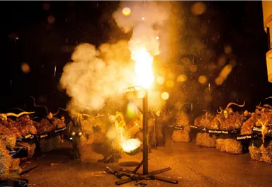 A festive scene with people in lively clothing gathered around a fire. Smoke and lights create an exciting atmosphere in the darkness.