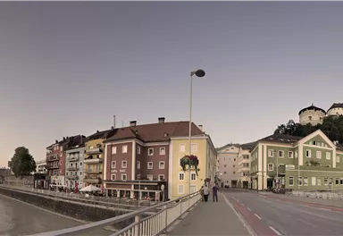 Eine malerische Uferpromenade mit bunten Häusern und einem Fluss. Im Hintergrund erhebt sich ein Hügel mit einer Burg.