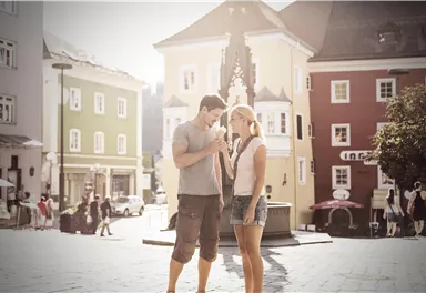 Ein Paar steht in einer sonnigen Stadtmitte und genießt Eis. Im Hintergrund sind bunte Gebäude und Passanten zu sehen.