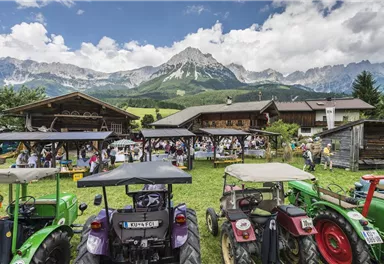 Eine ländliche Veranstaltung mit Traktoren im Vordergrund und festlichen Aktivitäten im Hintergrund. Die Berge sind majestätisch in der Ferne sichtbar.