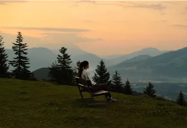 Eine Person sitzt alleine auf einer Bank und schaut auf eine malerische Berglandschaft während des Sonnenuntergangs. Die Szene ist von Bäumen umgeben und vermittelt eine ruhige, entspannende Atmosphäre.