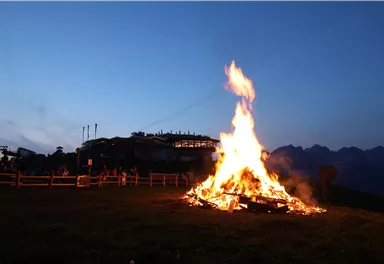 A large fire burns in a meadow at dusk. In the background, people and a building view are visible.