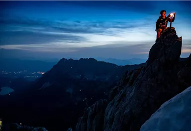 A mountain climber stands on a rock and holds a torch in their hand. The sky is dark blue, and the landscape is surrounded by mountains.