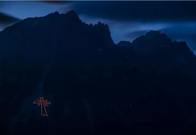 A large illuminated cross on a mountain in the dark. The surroundings are rough and mountainous, with dark clouds in the sky.