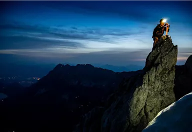 Ein Bergsteiger steht auf einem hohen Felsen und untersucht seine Ausrüstung. Im Hintergrund sind die bergigen Landschaften und ein klarer Himmel bei Dämmerung zu sehen.