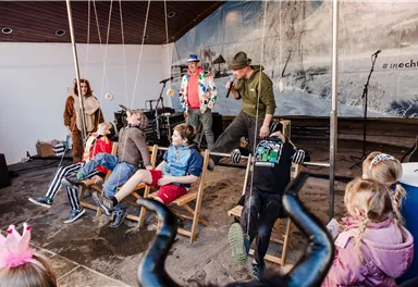 Eine fröhliche Veranstaltung mit Kindern und Erwachsenen, die an einer Bühnenshow teilnehmen. Die Kinder sitzen in Liegestühlen, während die Darsteller humorvolle Aktivitäten präsentieren.