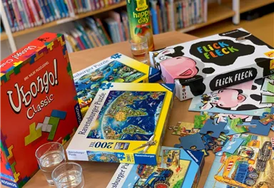 Ein Tisch mit Gesellschaftsspielen und Puzzles liegt in einer Bibliothek. Verschiedene Spieleboxen und Puzzlestücke sind sichtbar.