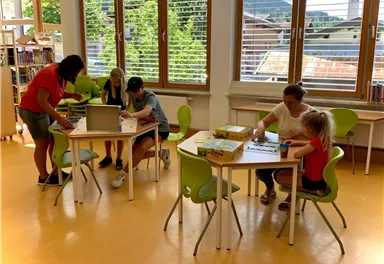 Eine Gruppe von Kindern arbeitet an Tischen in einem hellen Raum. Sie sind mit Spielen und Bastelmaterialien beschäftigt.