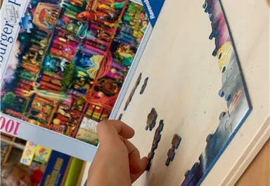 Ein Kind puzzelt an einem bunten Puzzle mit vielen Teilen. Im Hintergrund ist die Verpackung des Puzzles zu sehen.