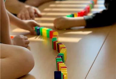 Zwei Kinder spielen auf einem Tisch mit bunten Dominosteinen. Sie bauen eine kreative Anordnung aus verschiedenen Farben.