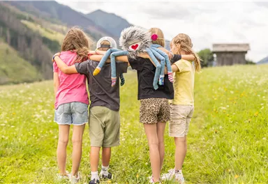 Vier Kinder stehen auf einer Wiese und umarmen sich. Eines der Kinder hält ein kuscheliges Spielzeug in Form eines Monsters.