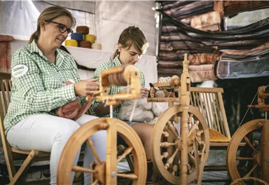 Zwei Frauen sitzen an Spinnrädern und arbeiten mit Wolle. Sie tragen kariert gestreifte Blusen und konzentrieren sich auf ihre Handarbeit.