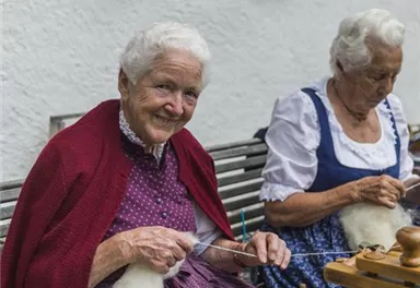 Zwei ältere Frauen sitzen auf einer Bank und handarbeiten mit Wolle. Sie tragen traditionelle Trachten und lächeln freundlich.