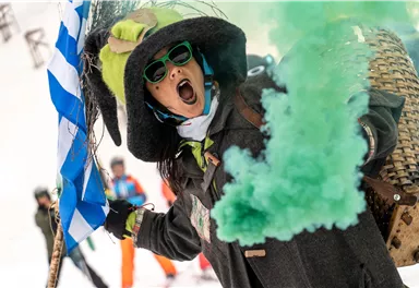 Eine Person trägt einen großen Hut und Sonnenbrille und feuert eine grüne Rauchfackel ab. Im Hintergrund sind Menschen auf Ski zu sehen.