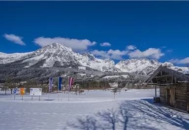 A snow-covered landscape with majestic mountains and a clear blue sky. In the foreground stands a wooden building and colorful flags are set up.