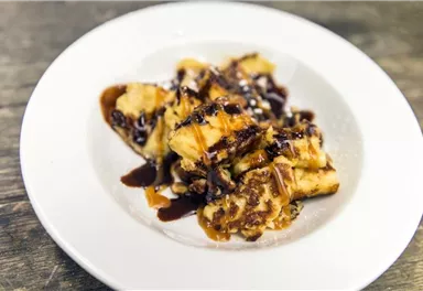 Ein köstliches Dessert auf einem weißen Teller. Es besteht aus frittierten Bananen, garniert mit Schokoladensauce und Puderzucker.