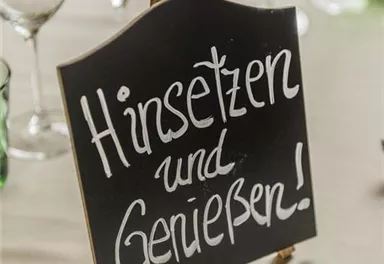 Ein kleines Schild mit der Aufschrift „Hinsetzen und Genießen!“ steht auf einem Tisch. Im Hintergrund sind Gläser und Kerzen sichtbar.