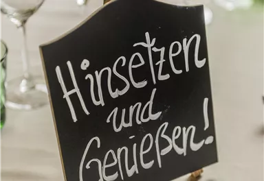 Eine Tafel mit der Aufschrift „Hinsetzen und Genießen!“ steht auf einem Tisch. Im Hintergrund sind Gläser und bunte Glasbehälter zu sehen.